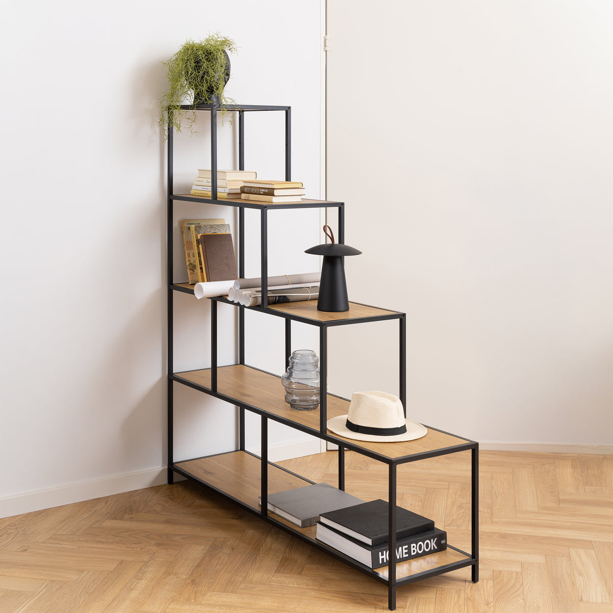 Skeleto-boekenkast 135x150, Wild Eiken, in de hoek van de kamer in visgraatpatroon, met boeken, een lampje en een hoed – trapvormige boekenkast