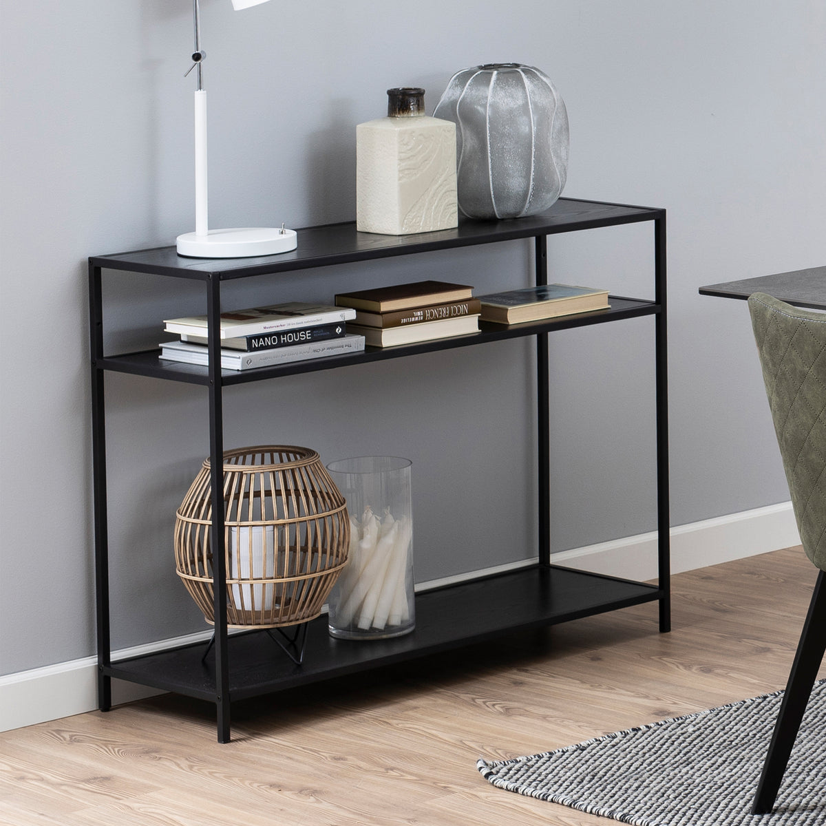 Console Skeleto 100 x 79 en mélaminé noir, avec des livres, une lampe et une lanterne sur les étagères, à côté d'une chaise près de la table
