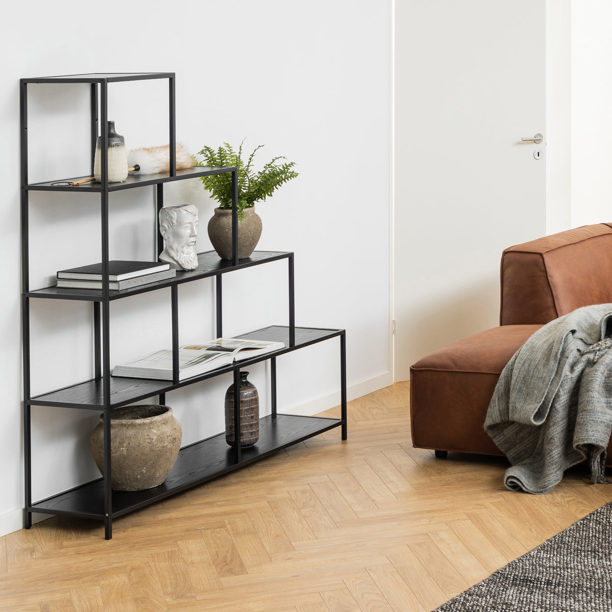 De Skeleto-boekenkast 135x150 in zwarte melamine in de woonkamer naast een bruine bank, met een plant, een buste en boeken