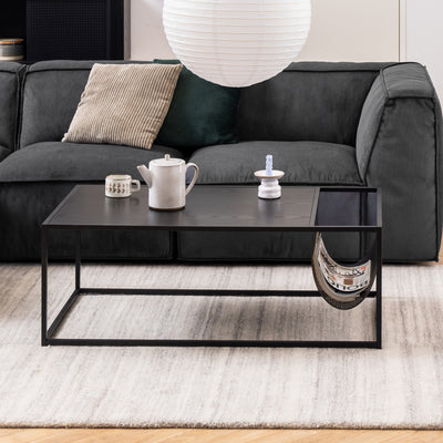 Table basse Skeleto 110 x 60 devant un canapé graphite posé sur un tapis clair, avec une carafe et des magazines dans une pochette métallique